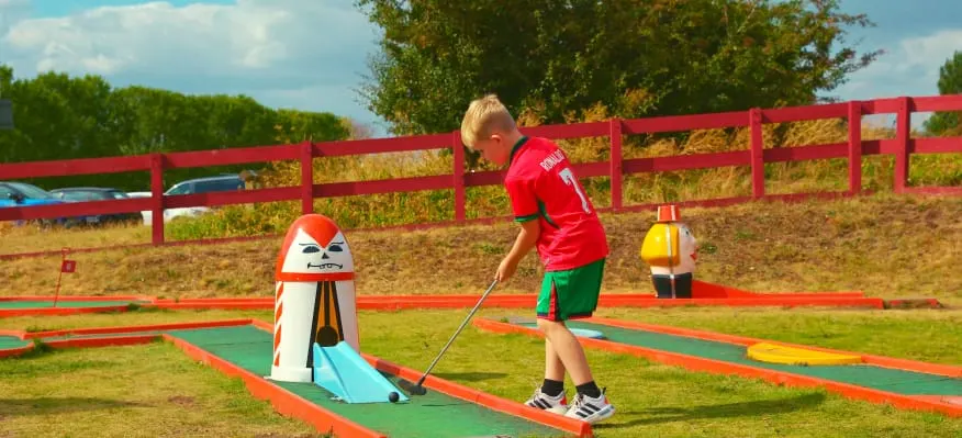 Child playing mini golf on a coloured course with cartoon-themed obstacles and red fencing surrounding a field.