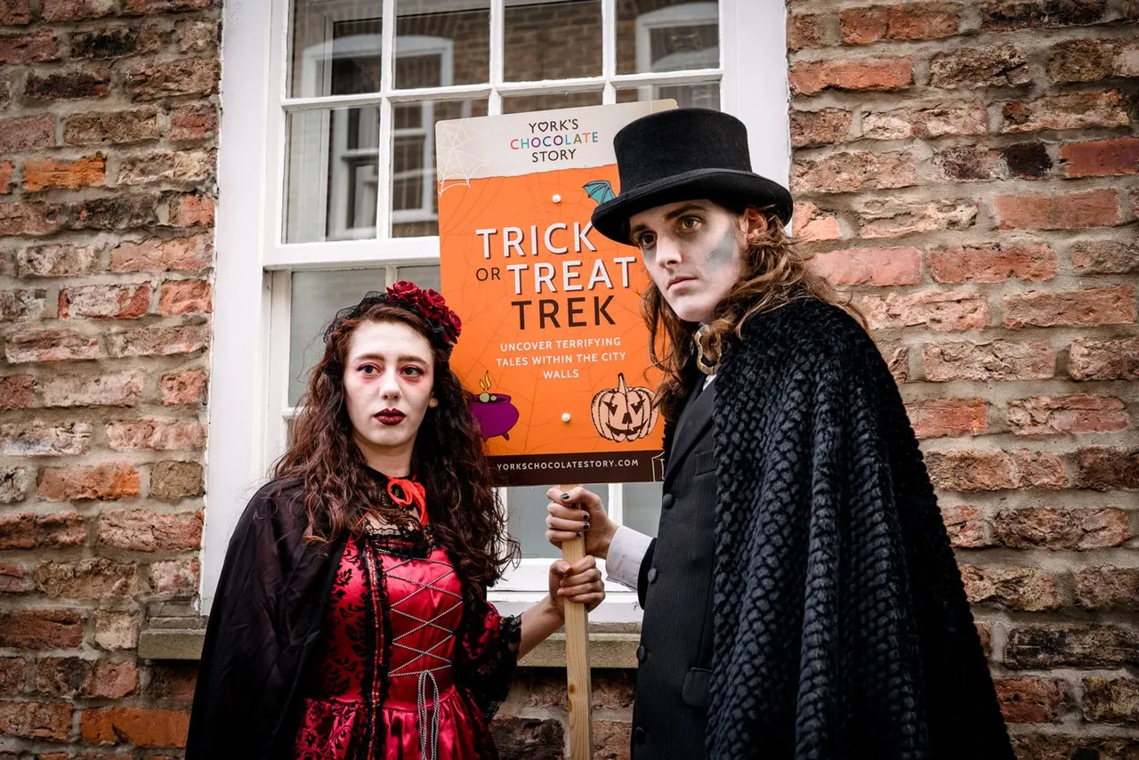 Two people in gothic Halloween costumes standing against a brick wall holding a "Trick or Treat Trek" poster.