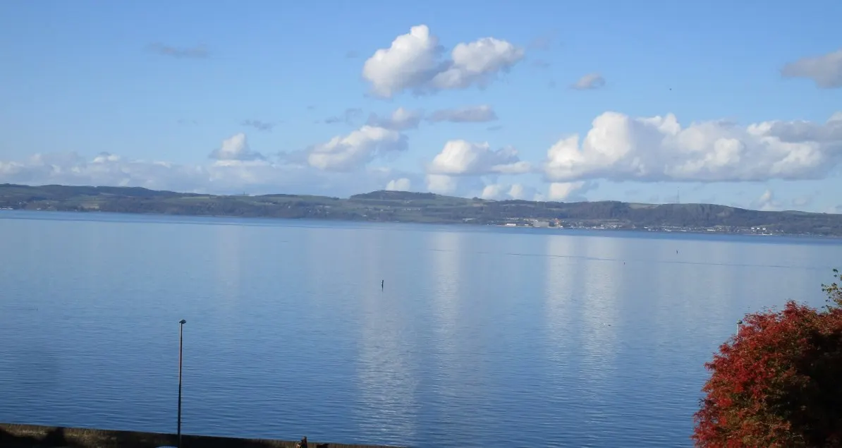 Calm blue loch with forested hills beyond under a partly cloudy sky, viewed from the shore.