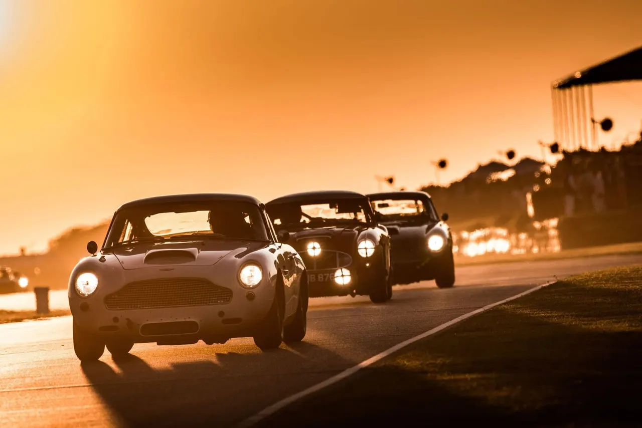 Classic sports cars driving in convoy on a circuit at golden hour with grandstand visible.