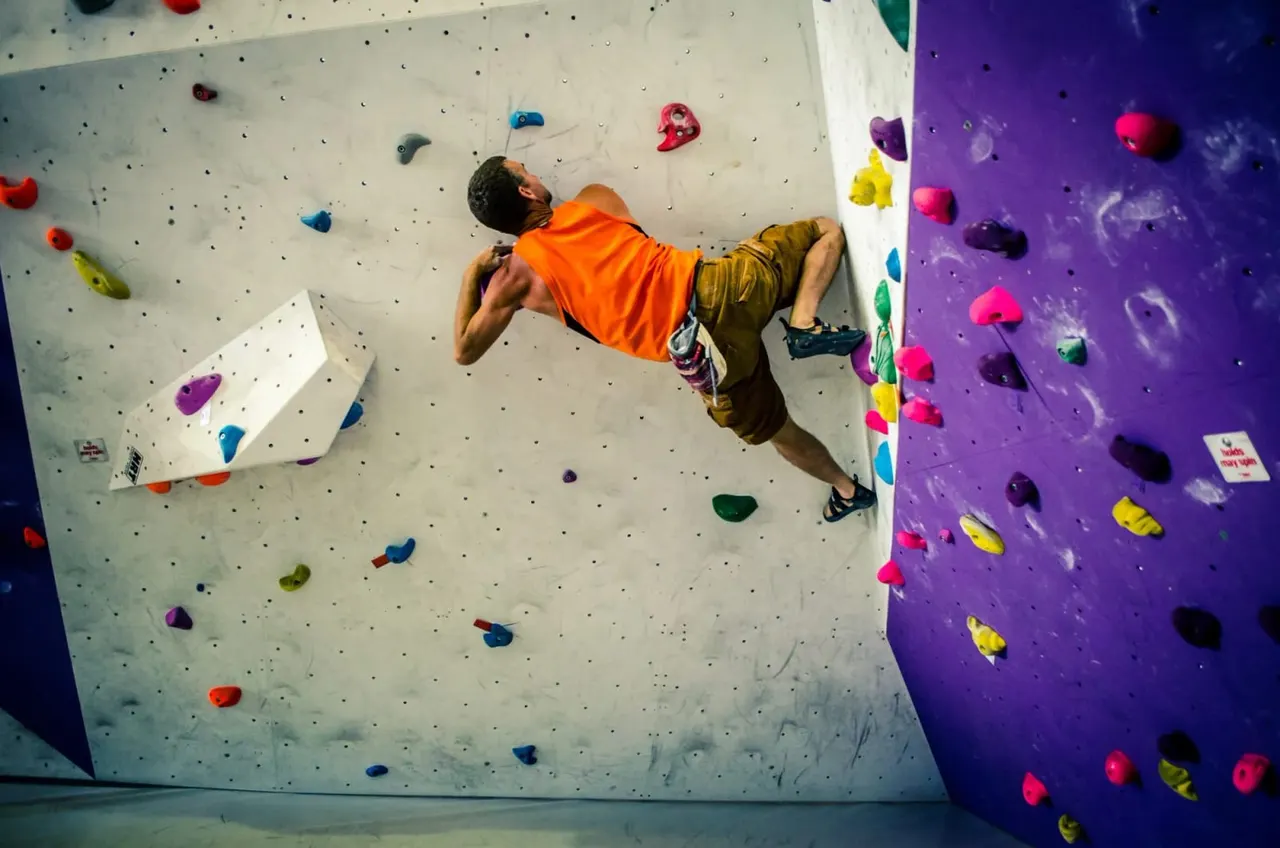 Man climbing indoor rock wall with coloured holds, wearing orange shirt and safety harness.