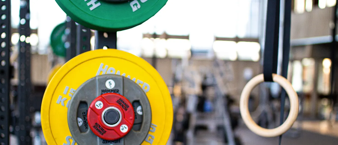 Yellow and red weight plates loaded on a barbell with gymnastics rings visible in a gym facility.