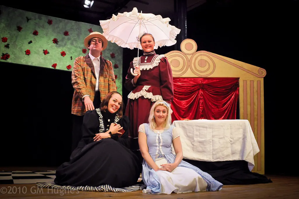 Four performers in period costume on a theatre stage during a comedic scene with ornate gold furniture and floral backdrop.