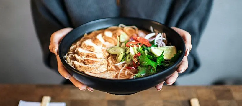 Person in dark top holding a black bowl of noodle soup with sliced meat and fresh vegetables.