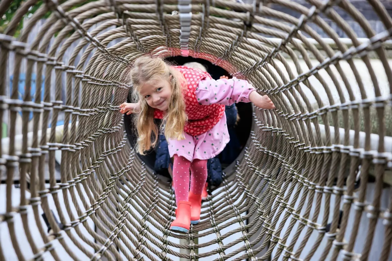 Young child crawling through a woven willow tunnel in a garden play area.