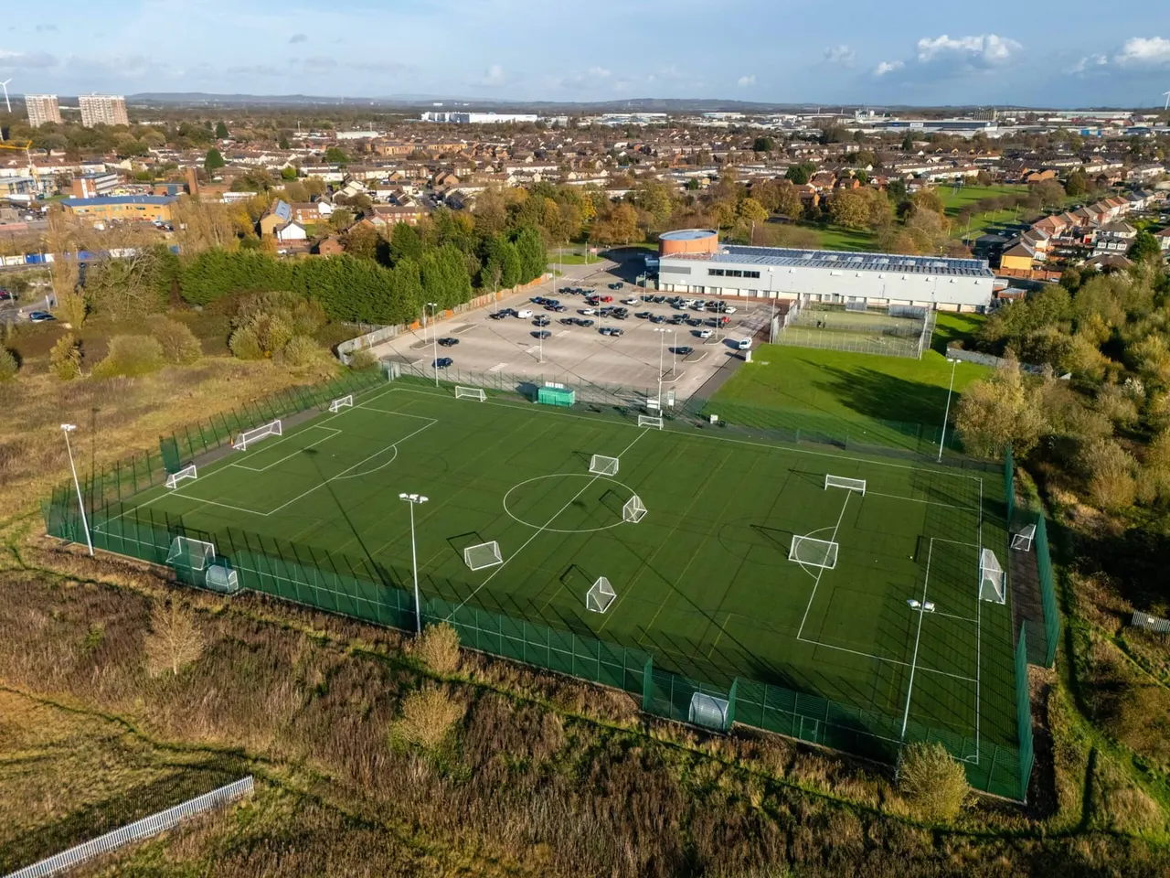 Aerial view of multiple football pitches with goals, surrounded by trees, a sports building with orange roof, and residential suburb in background.