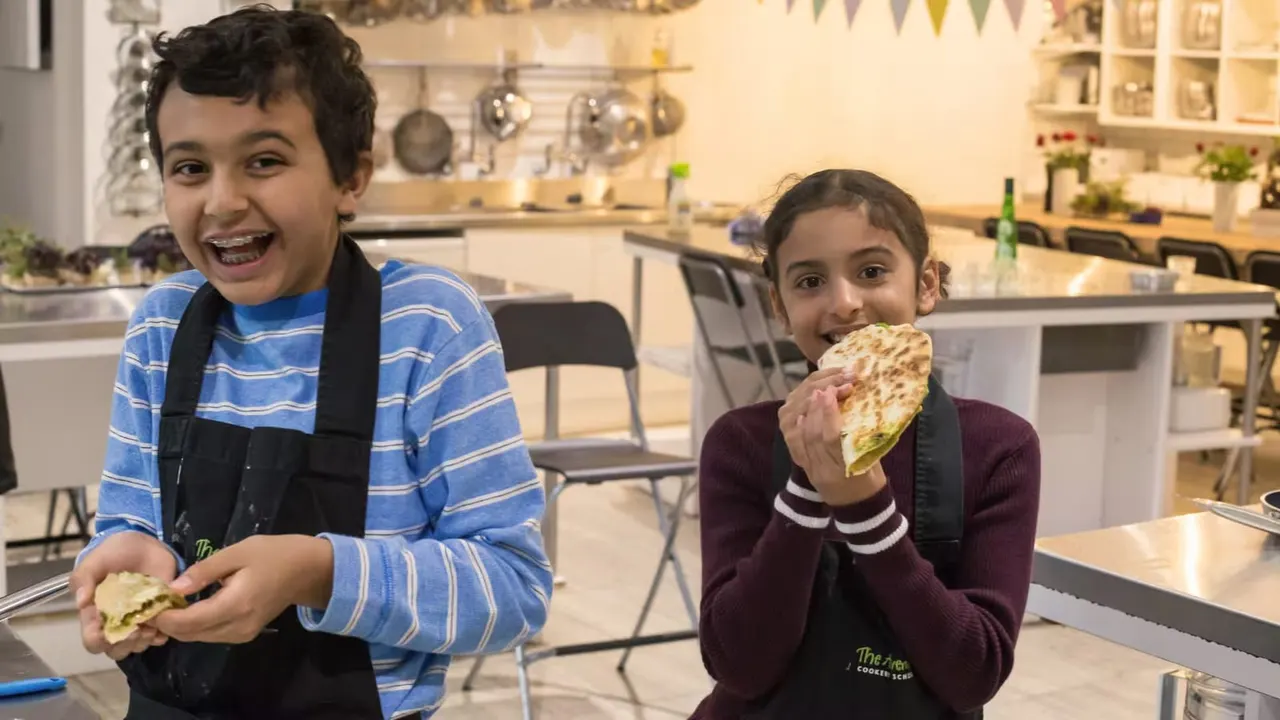 Two children in a cookery school kitchen holding and enjoying freshly made food.