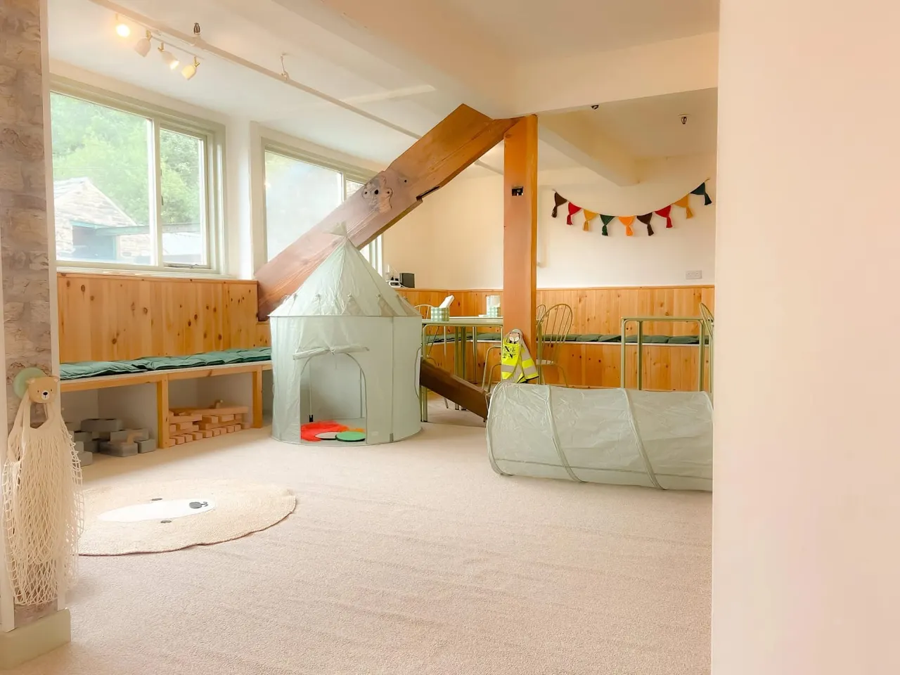 Bright indoor play room with wooden climbing structures, a slide, tent, tunnel, and raised platforms for children.