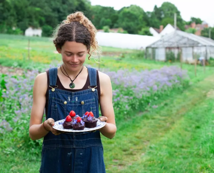 Woman in denim overalls holding a plate of chocolate cakes with raspberries in a farm field with lavender and greenhouse.