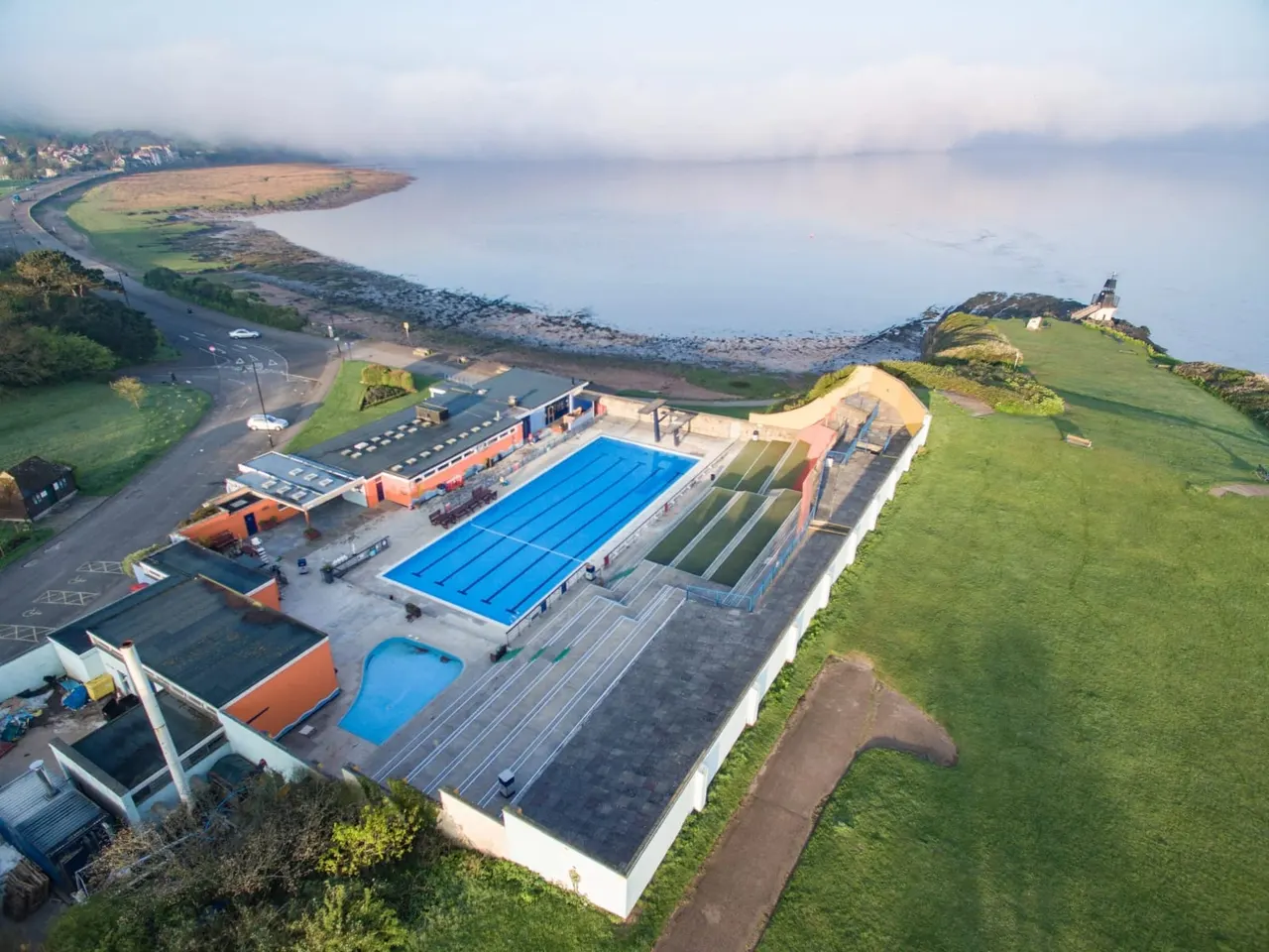 Aerial view of an open-air swimming leisure complex with blue pools, buildings and grounds beside a coastal estuary.