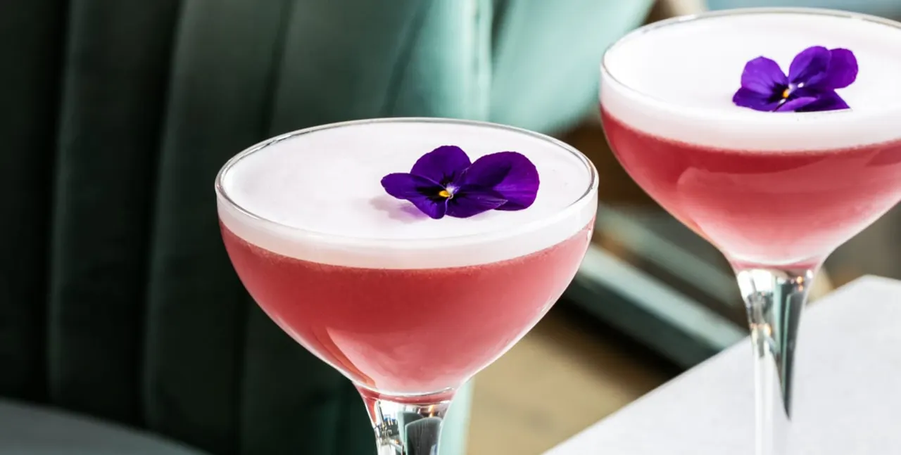 Two cocktails with pink liquid, white foam tops, and purple flower garnishes.