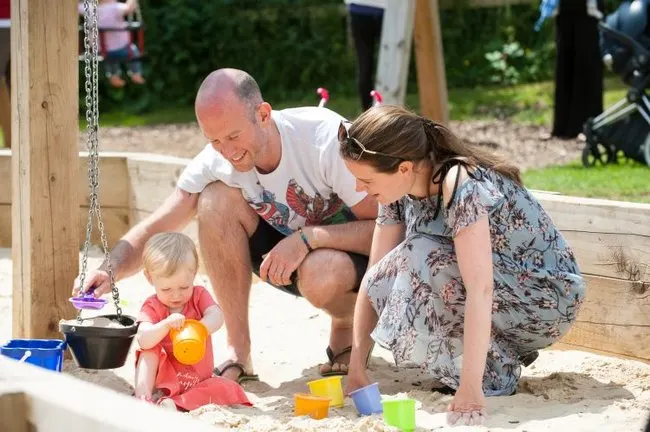 Man and woman crouch beside toddler playing with toys in sand pit.