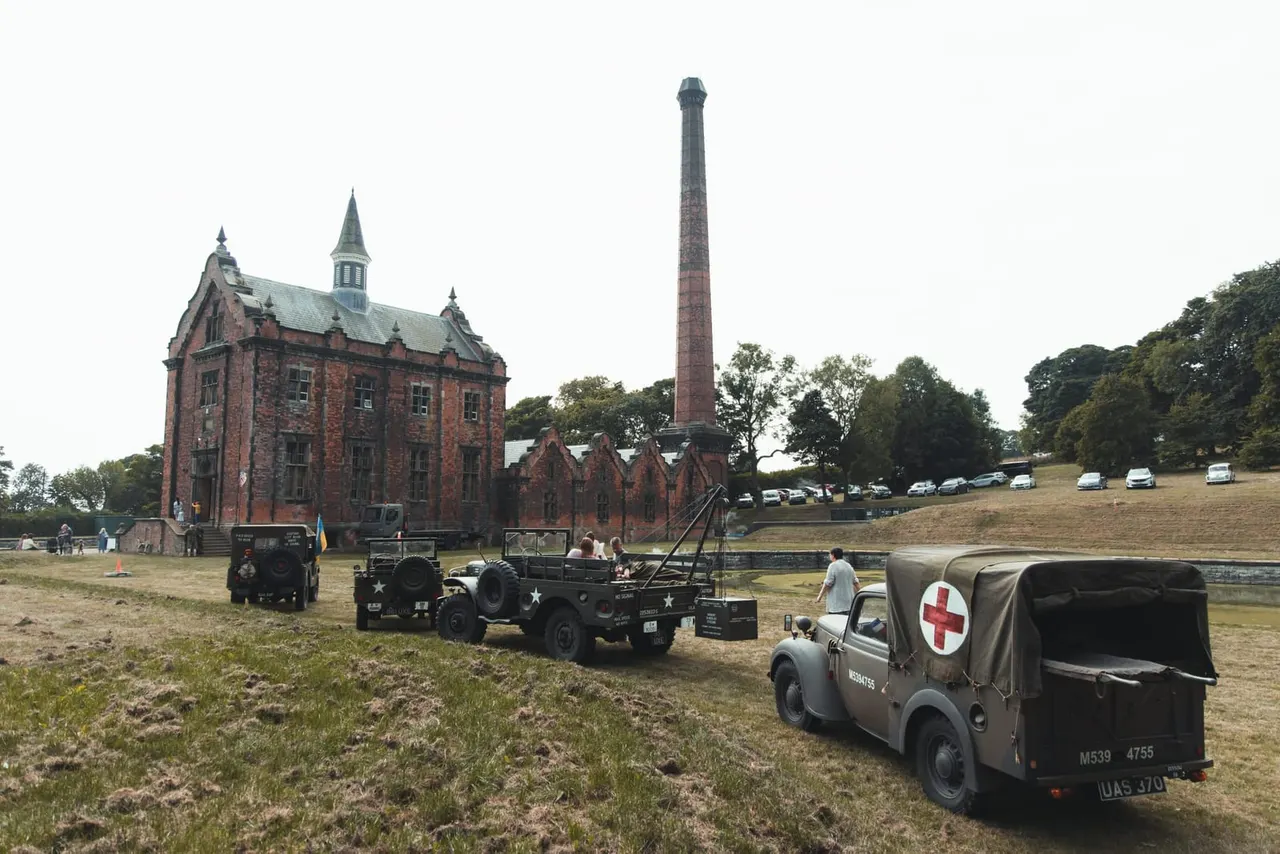 Vintage military vehicles and period ambulance parked on grass beside Victorian brick building and tall industrial chimney.