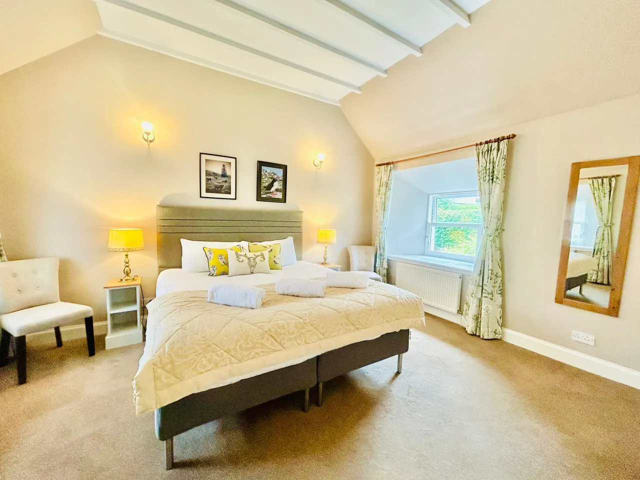 Spacious bedroom with cream and yellow décor, featuring a double bed with white pillows, seating area, and views over countryside.