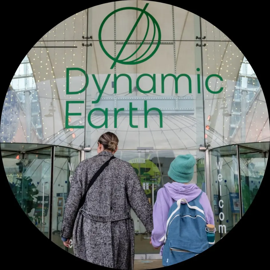 Two visitors approaching the entrance to a domed greenhouse building with the "Dynamic Earth" logo displayed.
