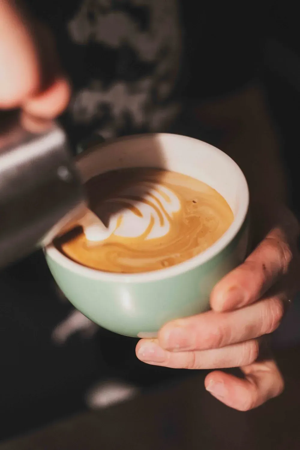 Barista pouring latte art into a mint-green cup held in someone's hands.