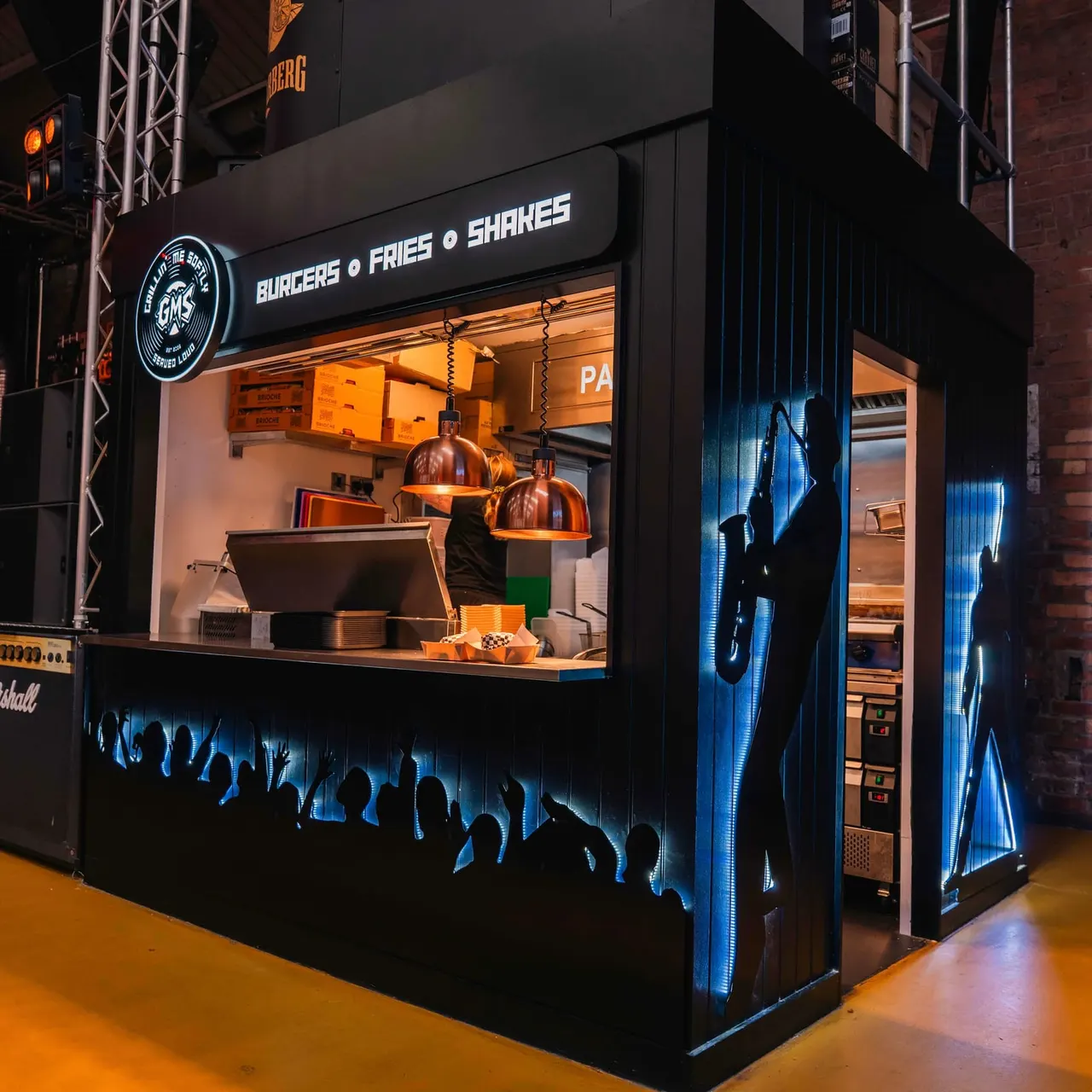 Fast food kiosk with neon signage serving burgers, fries and shakes, lit in orange and blue at night.