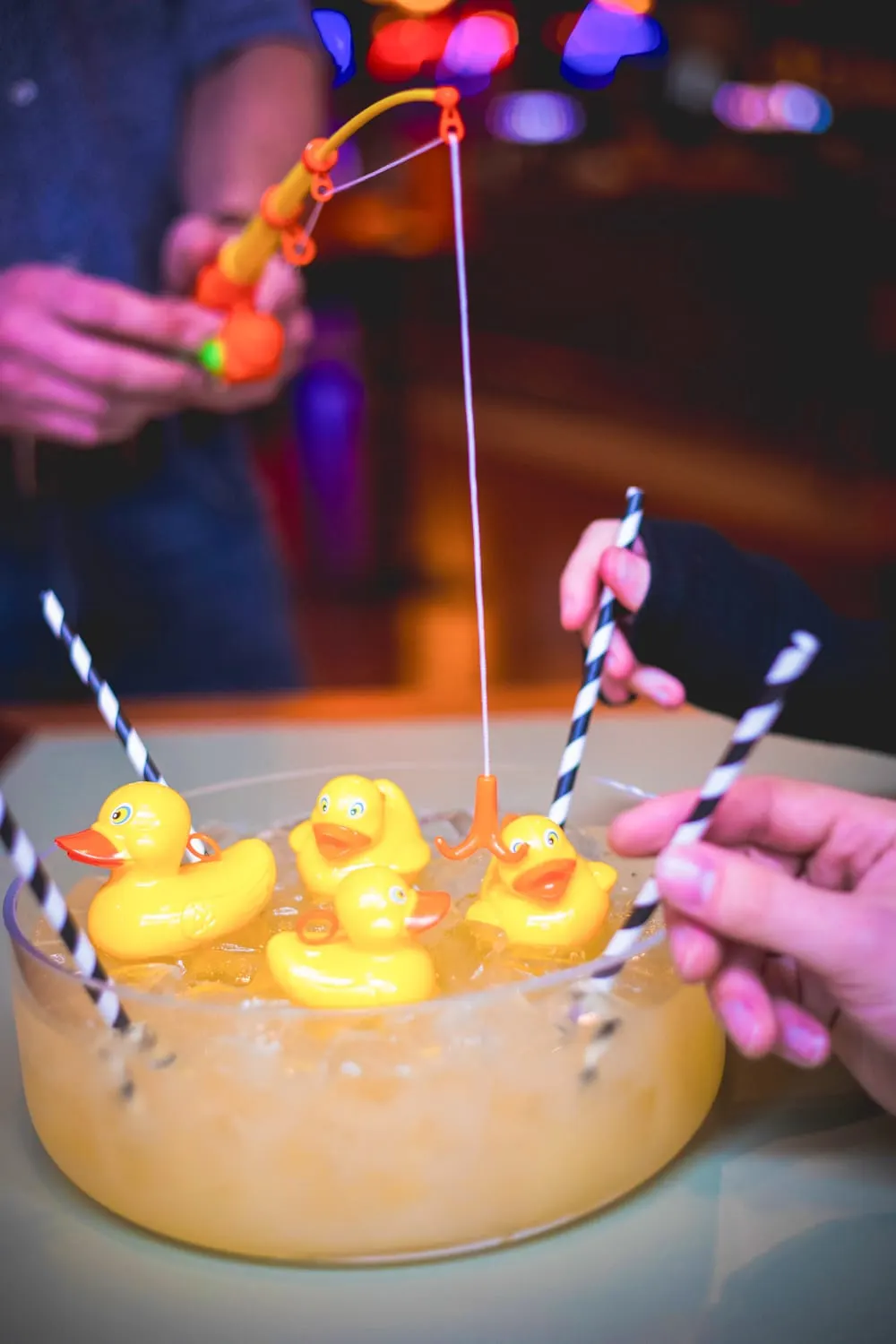Hand holding a fishing rod game above a yellow cake decorated with rubber ducks and striped straws, surrounded by colourful neon lights.