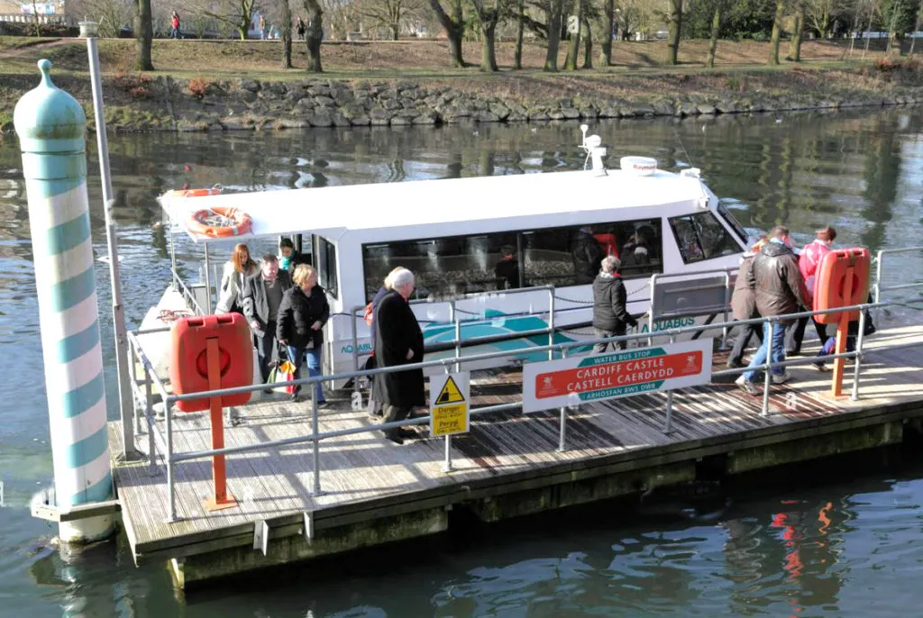 Passengers boarding a white river cruise boat at a wooden jetty on an urban waterway, with bare trees and parkland visible beyond.