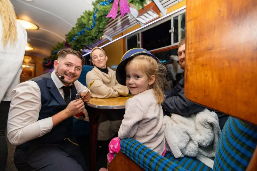 Staff member serves festive food to a young child inside a decorated vintage railway carriage with other passengers.