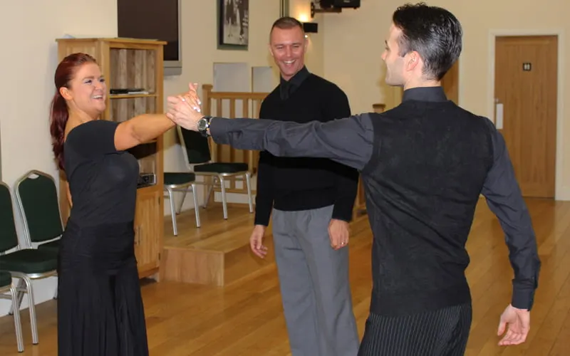 Three people in dark clothing performing a dance move with hands joined together in a wooden-floored studio.