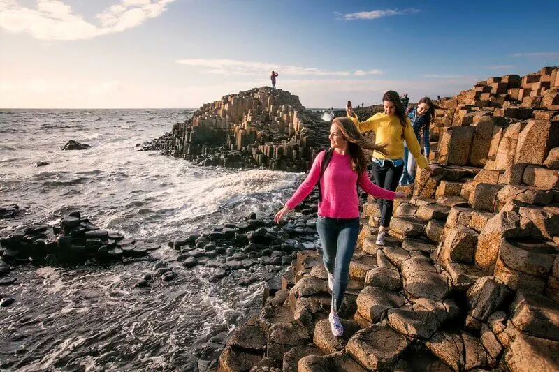 Visitors exploring hexagonal basalt columns at a rocky coastal headland beside the sea.