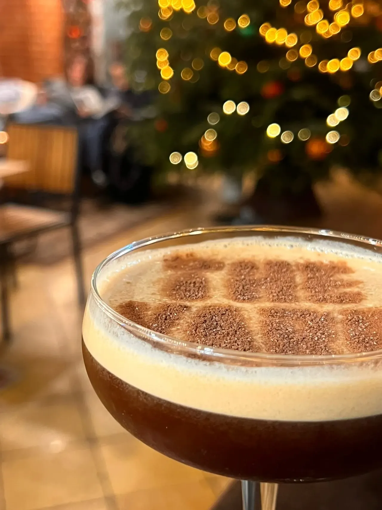 Tiramisu cocktail with cocoa powder dusted on top, served in a coupe glass with festive golden lights and blurred venue in background.