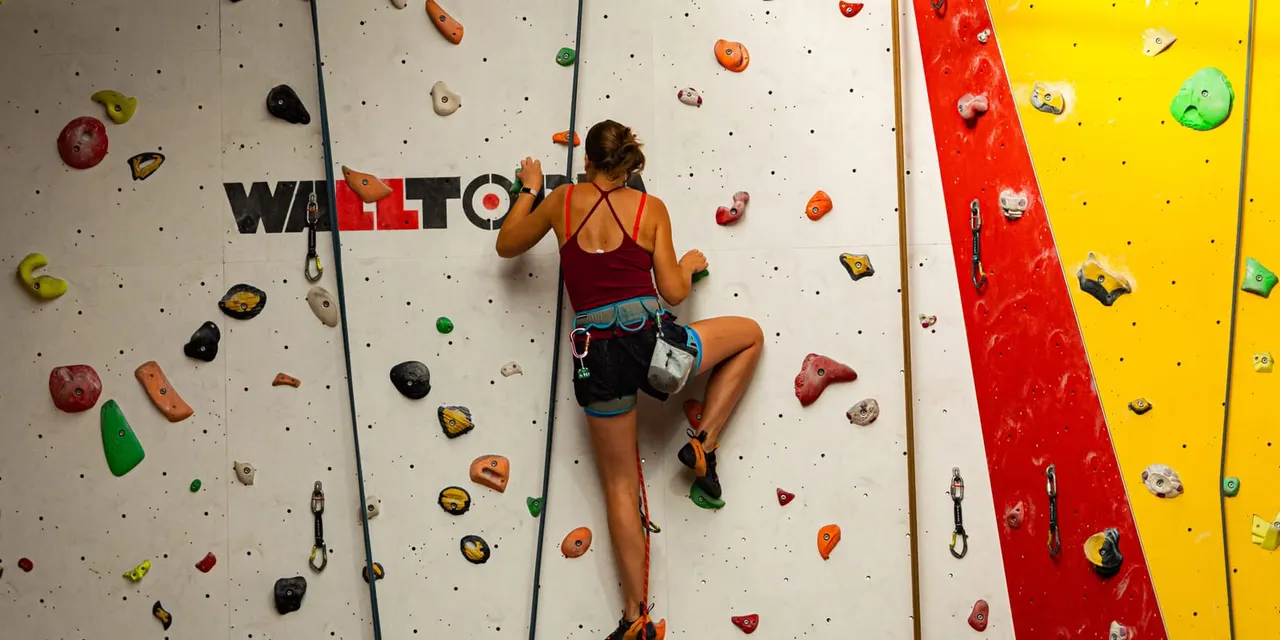 A climber ascending an indoor climbing wall with coloured holds, wearing safety equipment and harness.