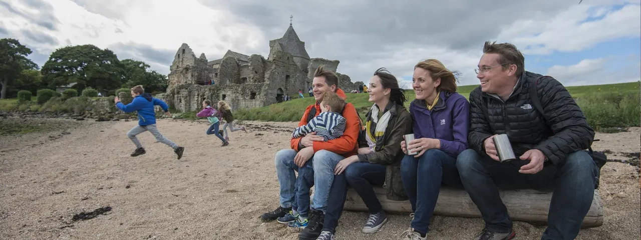 Family group sitting on a log overlooking a ruined castle on a beach, with children playing in the sand behind them.