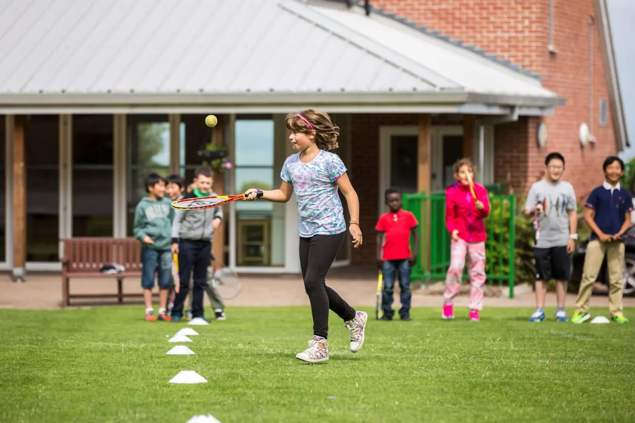 Girl playing tennis on a lawn whilst other children watch from behind at a brick building venue.