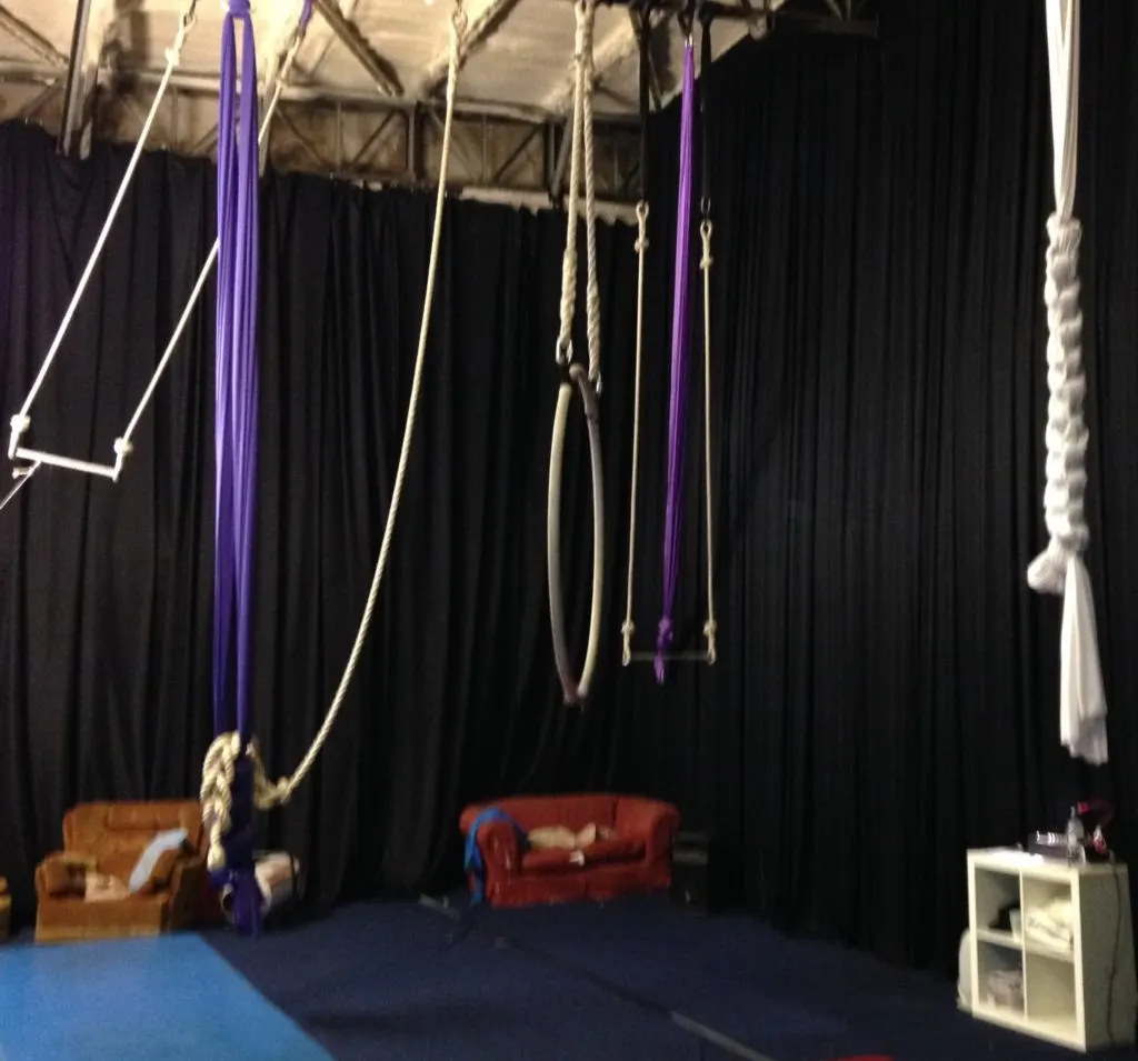 Aerial silks and ropes hang from a high ceiling in a circus training studio with black drapes and padded floor.