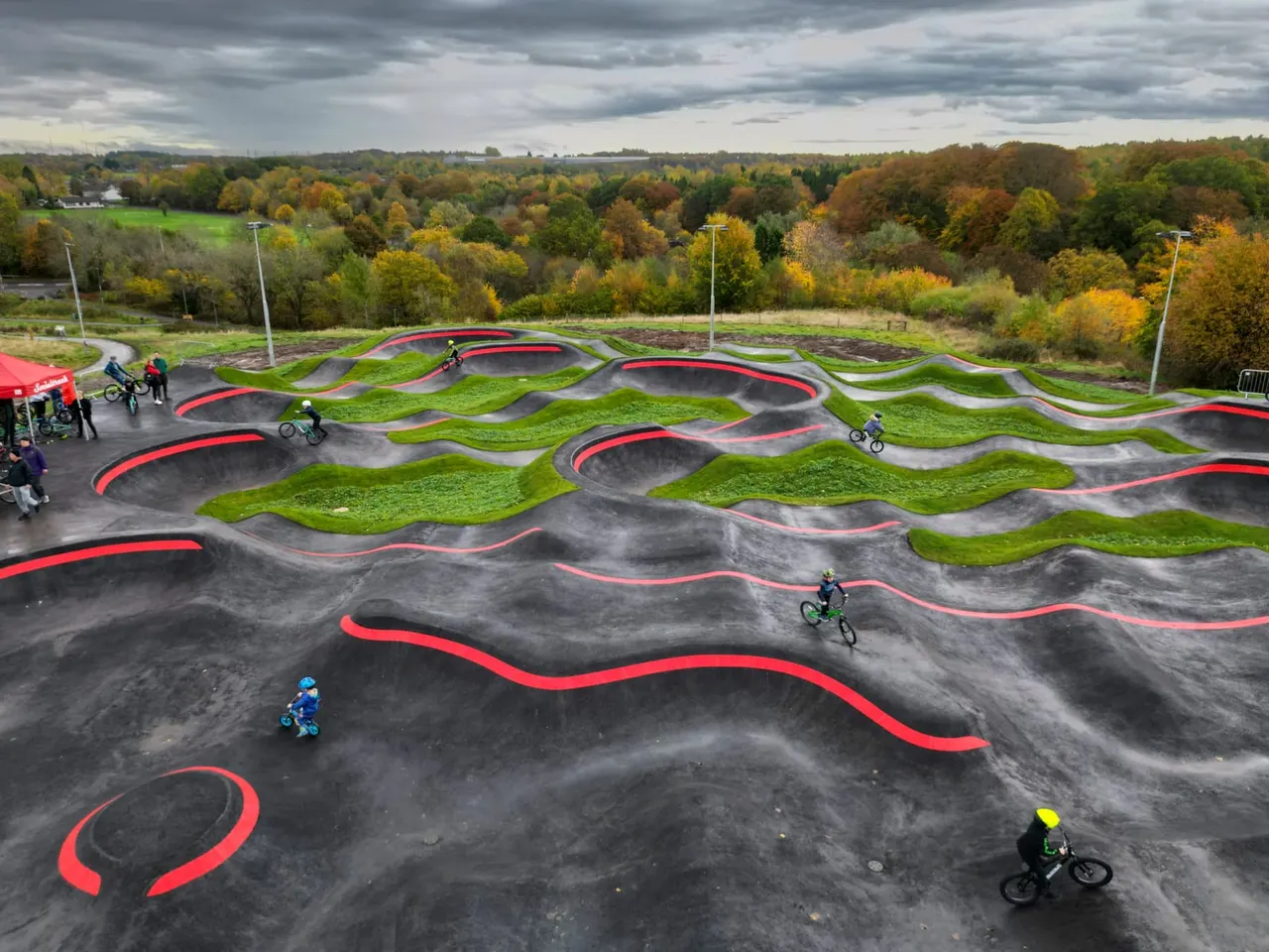 Multiple cyclists riding on a modern BMX track with red-edged black asphalt ramps set against autumn woodland landscape.