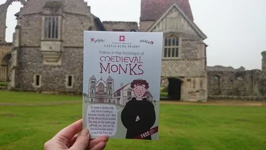 A visitor holds a leaflet about medieval monks in front of stone priory ruins and buildings.
