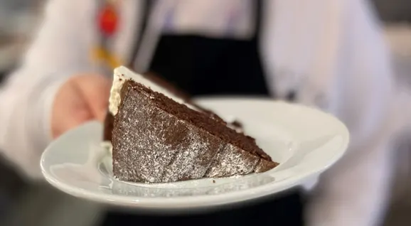 A server in white chef's uniform holds a triangular slice of chocolate cake dusted with icing sugar.