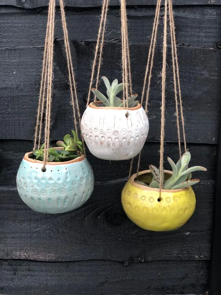 Three hanging planters with succulents in white, pale blue and yellow ceramic pots against a dark wooden wall.