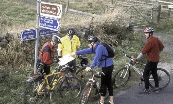 Group of four cyclists in colourful jackets gathered at a countryside signpost to check their route.