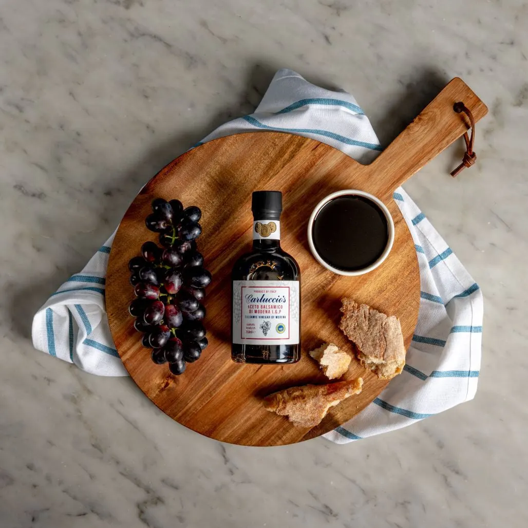 Wooden board with balsamic vinegar bottle, dark grapes, bread, bowl of vinegar and striped cloth on marble surface.