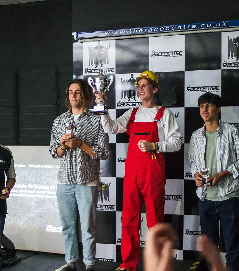 Three young men stand on a chequered podium holding trophies at a go-kart racing venue.