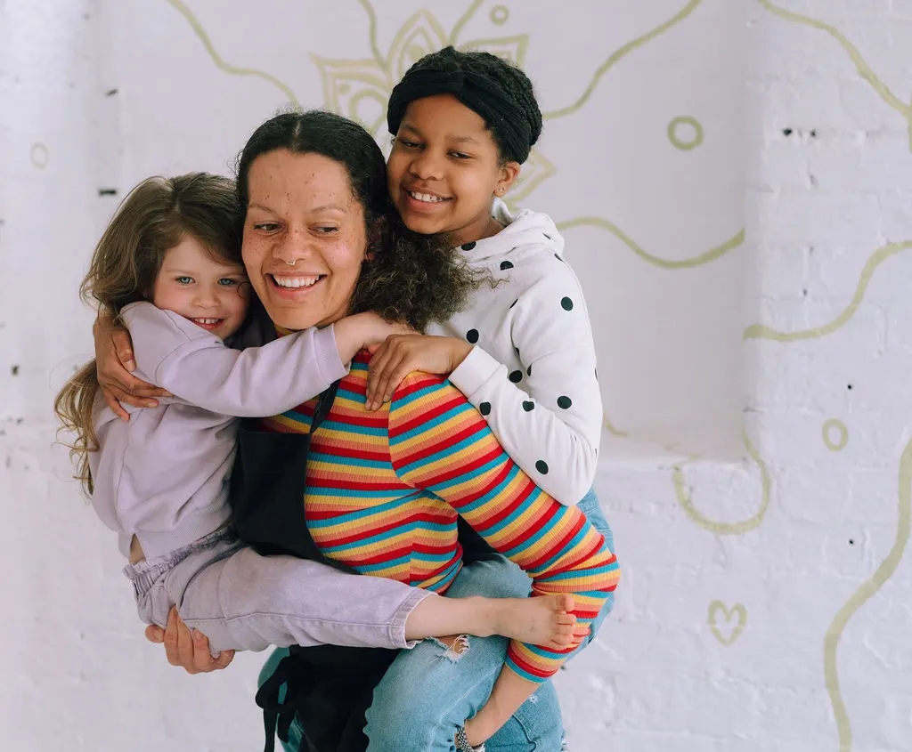 Woman holding two young children, smiling at camera against a white wall with decorative patterns.