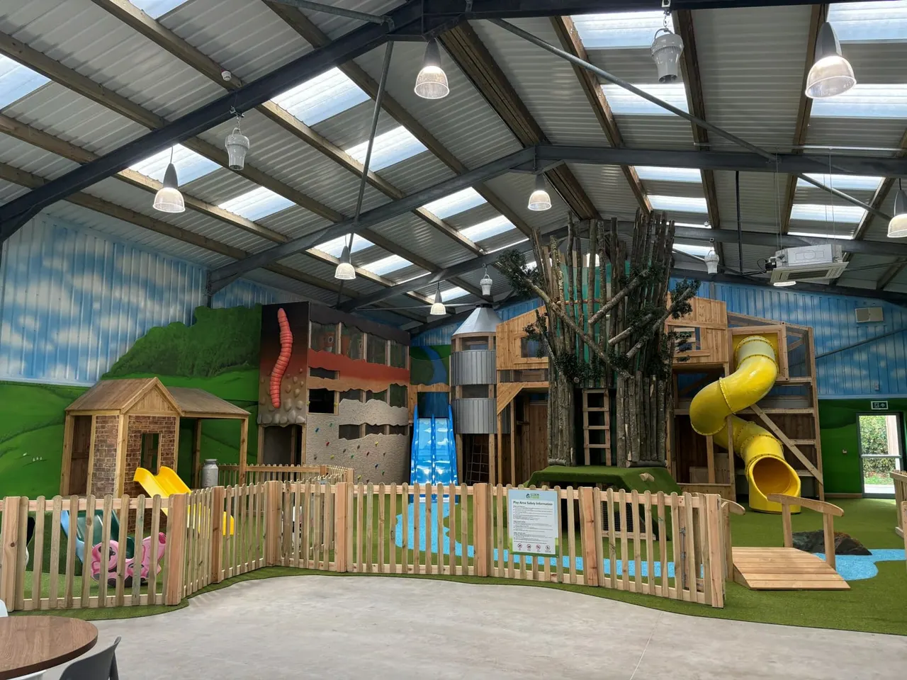 Indoor play area with wooden structures, slides, climbing frames and a worm tunnel feature enclosed within a corrugated steel building.
