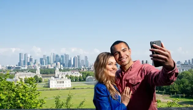 Couple taking a selfie with a city skyline and historic building visible behind them.