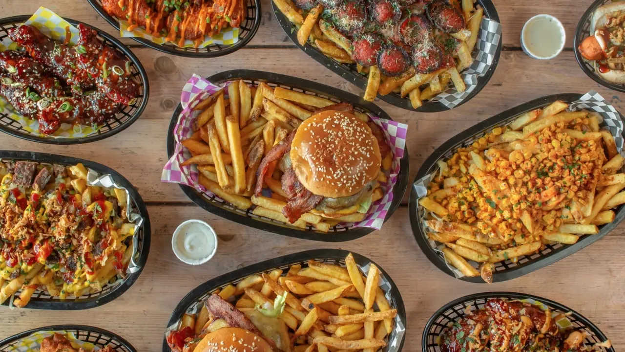 Overhead view of fast food meal spread including burgers with fries, loaded fries with toppings, and pizza on wooden table.
