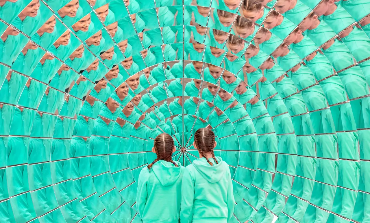 Two children stand inside a tunnel of mirrors reflecting their images repeatedly in turquoise light.