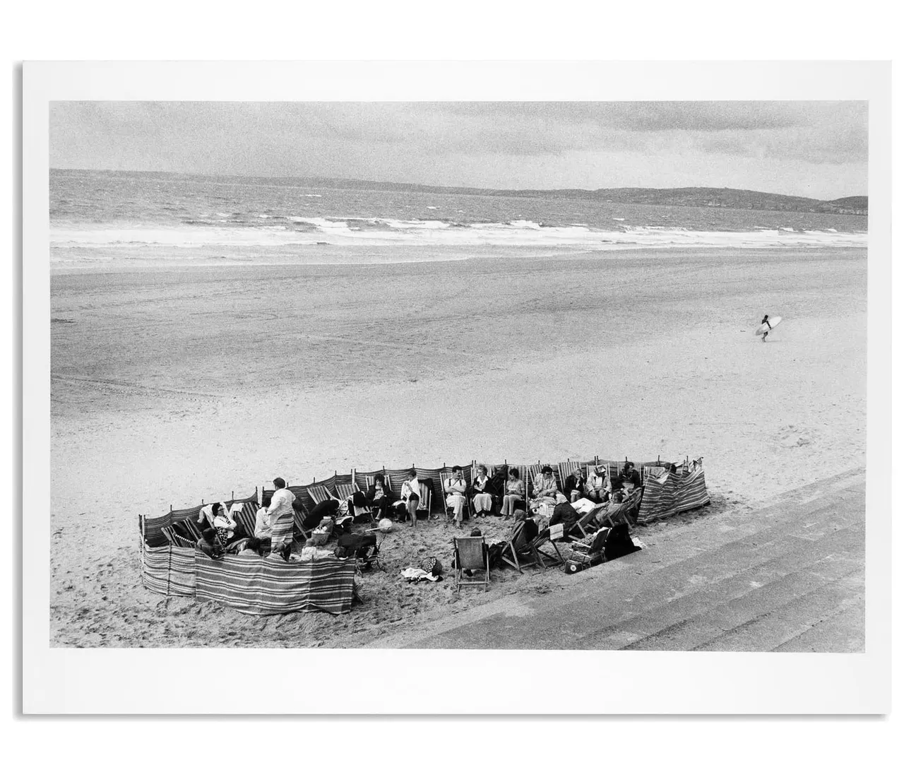 Group of beachgoers sitting in striped deckchairs sheltered by canvas windbreaks on a sandy beach with waves and hills beyond.