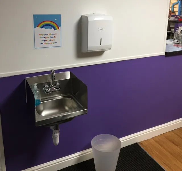 Stainless steel hand-washing sink mounted on purple wall with paper towel dispenser and hygiene poster above.