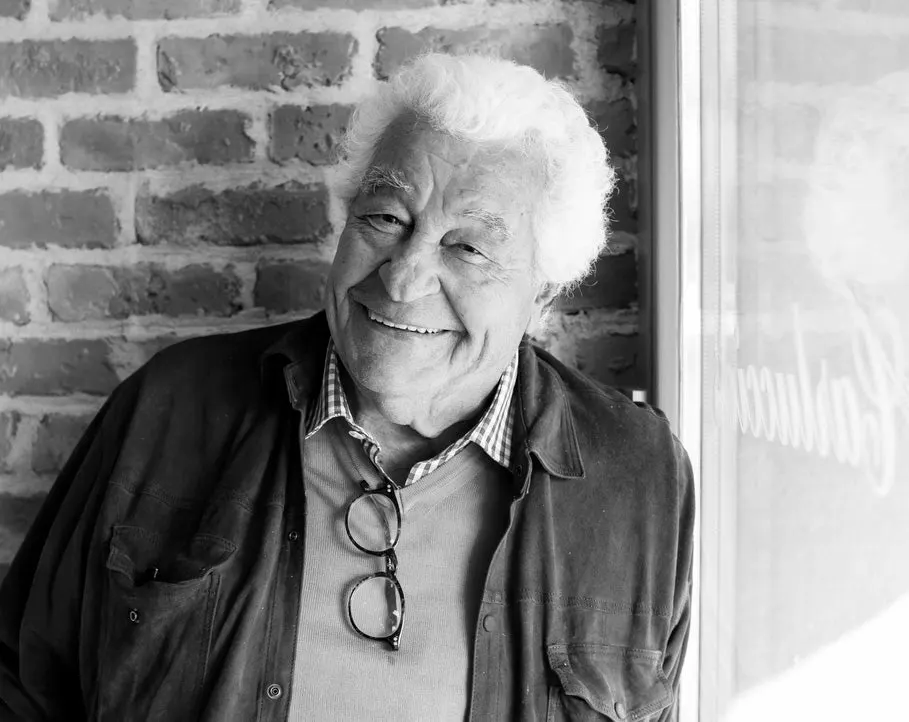 An older man with white hair smiles at the camera against a brick wall.