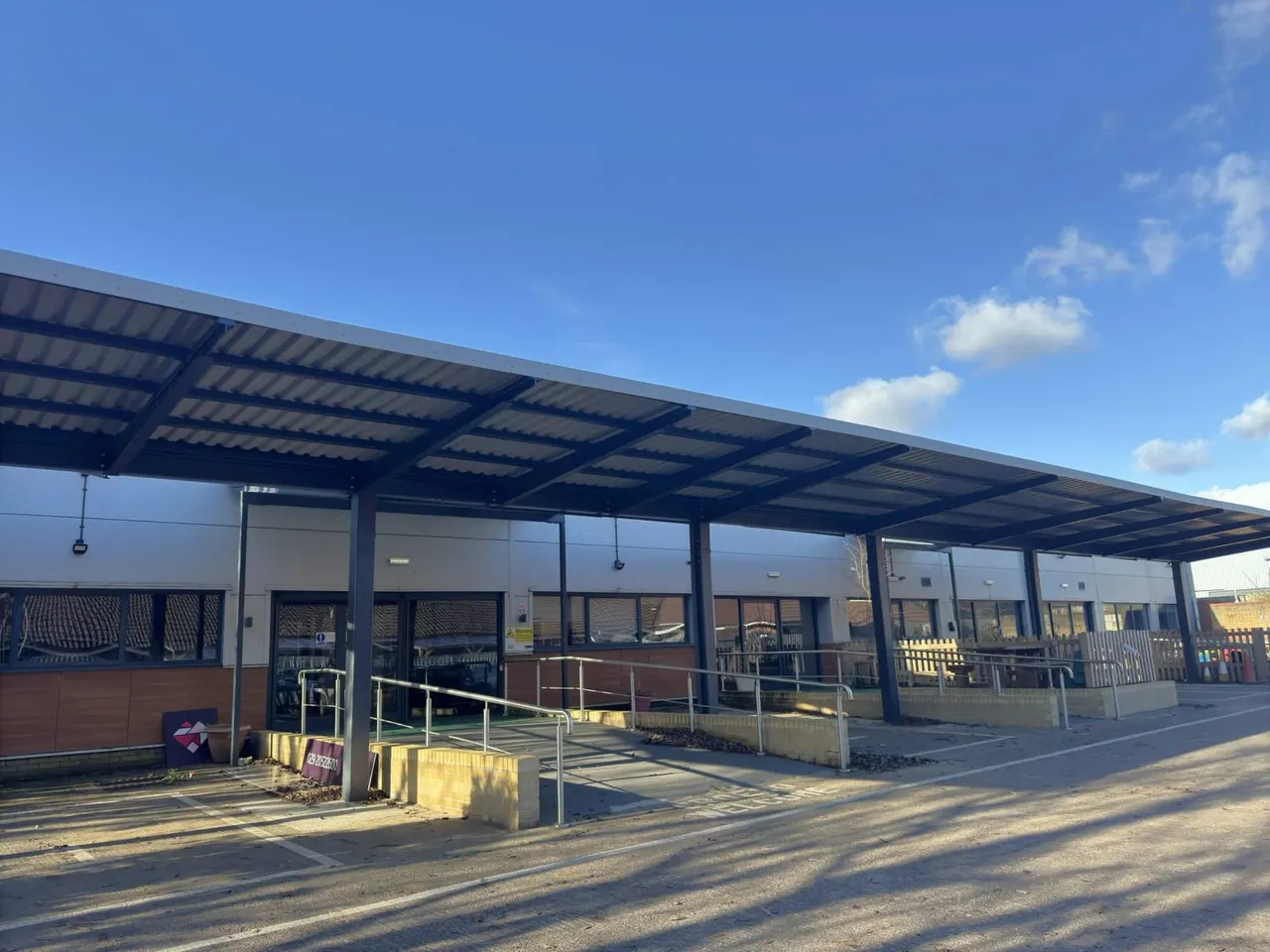Modern single-storey building with metal roof canopy, large windows, and metal railings under clear blue sky.