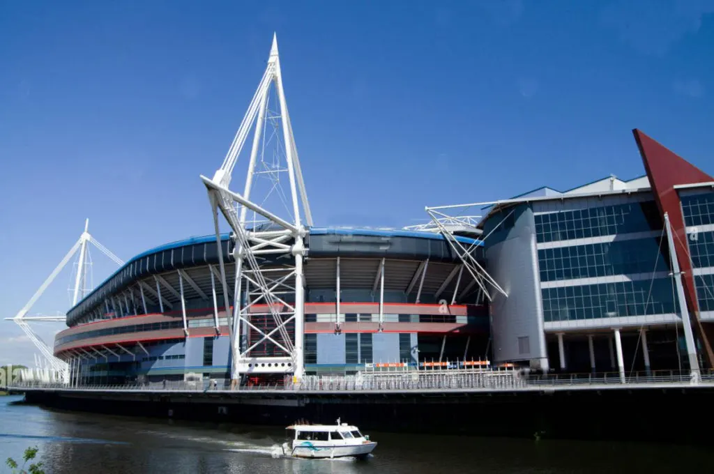 Modern waterside sports stadium with white cable-stayed structures, viewed from the water with a small white boat in foreground.