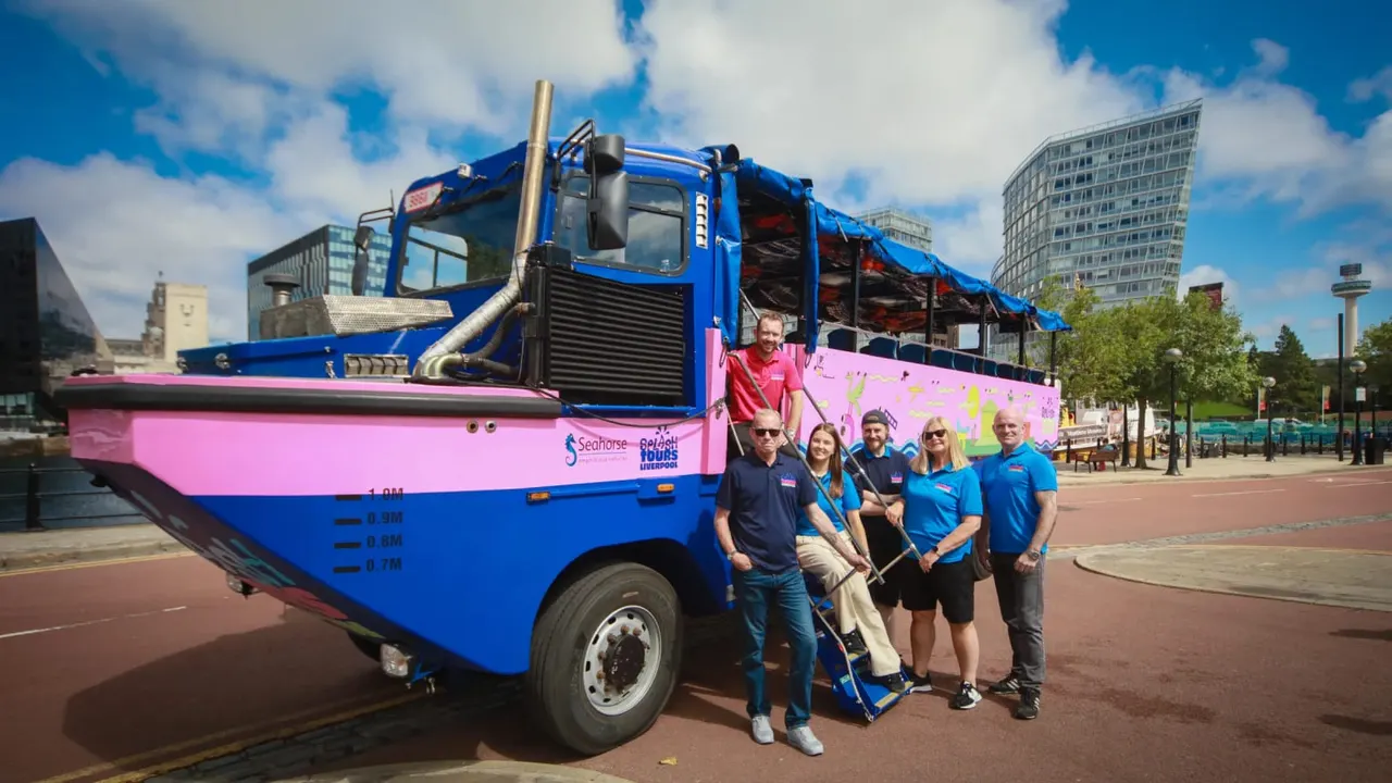 Group of staff members posing in front of a blue and pink amphibious tour vehicle in an urban waterfront setting.