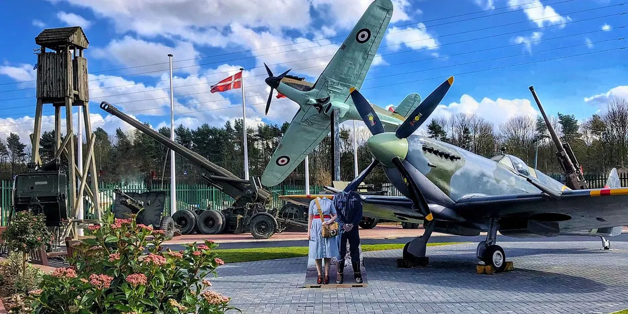 Two visitors standing before a restored RAF Spitfire aircraft displayed outdoors at a military museum, with vintage weaponry and buildings visible nearby.